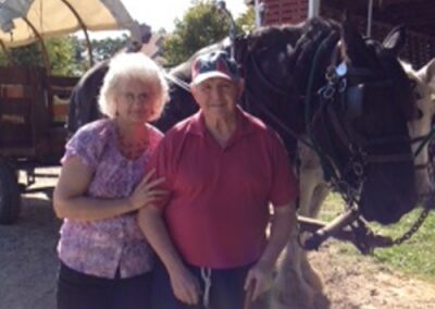 An elderly couple standing beside a black horse near a rustic barn.