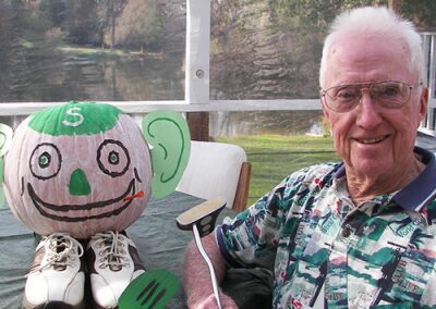 An elderly man smiles holding a pink pen next to a decorated pumpkin with shoes.